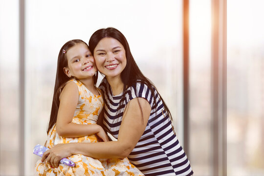 Portrait of a happy asian woman with her adopted caucasian little daughter. Cute little girl with her mother indoors. Blurred windows background.