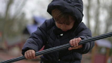 Toddler boy playing at playground gripping rope. Child wearing winter clothes plays outdoors holding cable