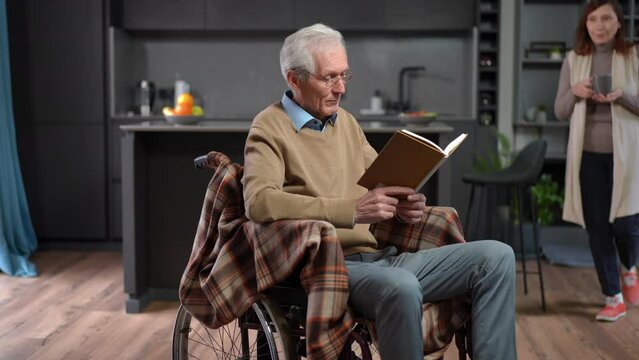 Portrait of intelligent senior man in wheelchair reading book as woman bringing coffee cup from kitchen. Handsome Caucasian husband enjoying hobby in the morning with wife at home