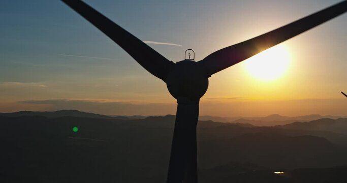 Aerial Cinematic Close-up Shot of Wind Turbines Rotating and Generating Green Ecological Energy. Windmill Producing Clean Renewable Energy. Background with Beautiful Mountains Landscape and Sunset