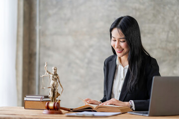 Asian female lawyer or legal advisor working with scales of justice Sitting at her desk holding a book on the scale of jurisprudence concepts to study and find information.