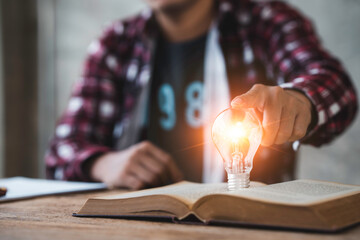 Young Asian businessman pointing at a light bulb placed on a book to generate ideas new ideas in management planning events Working in the modern age online world.