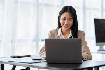 Asian young woman businesswoman using a laptop sitting smiling using modern analytical concepts While happily working at the desk in the office.