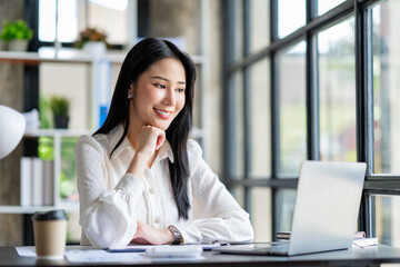 Portrait of a beautiful cheerful Asian businesswoman sitting at her desk with a cup of coffee while taking a break with a happy smile in her upcoming successful business.