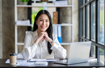 Portrait of a beautiful cheerful Asian businesswoman sitting at her desk with a cup of coffee while taking a break with a happy smile in her upcoming successful business.