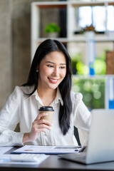 Portrait of a beautiful cheerful Asian businesswoman sitting at her desk with a cup of coffee while taking a break with a happy smile in her upcoming successful business.