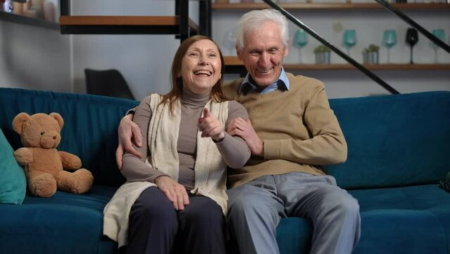 Front view portrait of relaxed senior couple hugging laughing watching comedy film on TV. Medium shot of carefree Caucasian handsome husband and beautiful wife enjoying leisure at home in living room