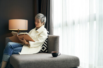 fashion Asian woman sitting in couch beside window reading