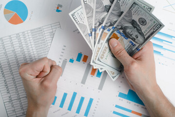 Close-up of hand holding money one hundred dollar bills on background of business documents with graphs and charts, top view. Concept of financial success, successful investment, wealth