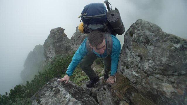 A man with a backpack climbs rocky mountains into the fog. A tourist conquers a mountain peak in the rain. A boy walks uphill next to large stones in the Carpathians. Concept: submission, fatigue. 4k