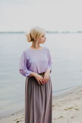 Blonde girl on a summer day on the shore near the river in nature in a dress