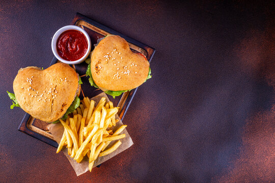 Valentine day Heart-shaped burgers. Two tasty cheeseburgers with french fries and beer bottles on dark table background. Idea for Valentine day dinner, dating, non-usual February 14 menu for couples