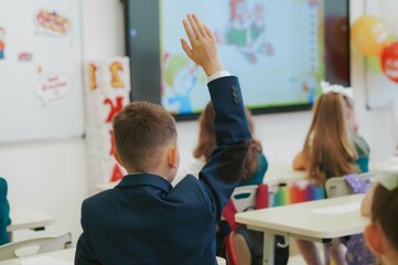 children in school uniforms sitting by desks. Boy raising hand to answer