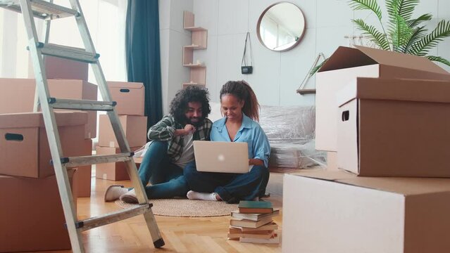 Young positive multiethnic African American woman and Arabian man interior designers working on residential renovations high five each other sits on floor with laptop in apartment with boxes