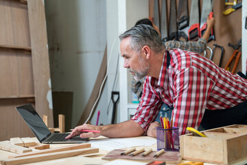 Middle-aged carpenter husband holding furniture blueprints for woodworking in laptop , making chairs, cabinets, interiors and carpentry as a hobby and doing furniture shop business.