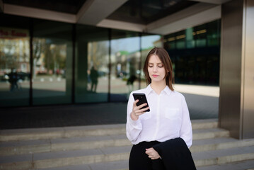 Fototapeta premium A business woman near an office building using a mobile phone answers a message on the Internet, communicates with business partners.