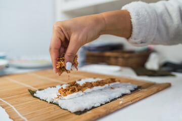 Unrecognizable woman's hands pouring and spreading some crispy onion on the nori seaweed sheet she is using to prepare homemade sushi in her kitchen.