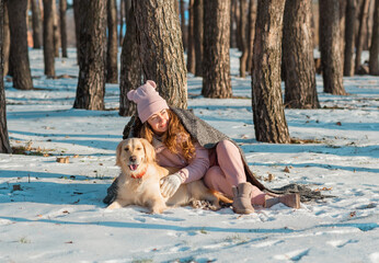 Portrait of a happy woman hugging her dog golden retriever covered together with a plaid on a snowy forest. Happy winter time concept.