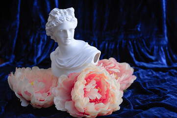 Greek sculpture among pink peonies on a blue velvet background. Copy space