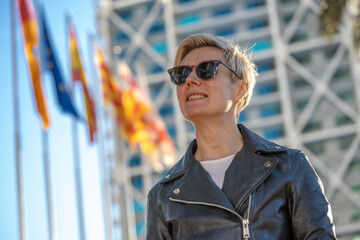 30s blond woman with short hair in sunglasses and leather jacket next to modern building in Barcelona with european flags on background