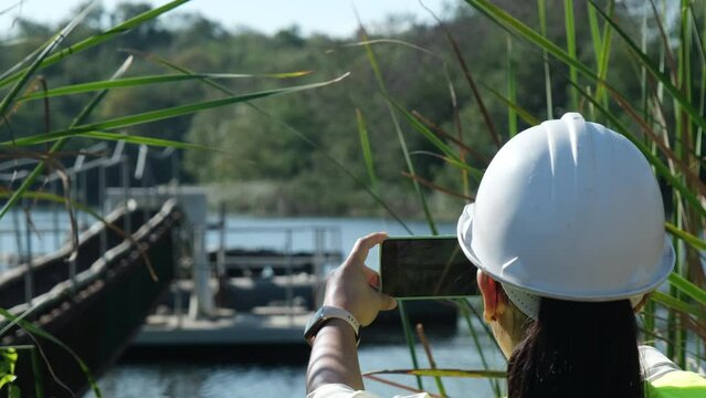 Environmental engineer wearing a white helmet uses a mobile phone to record data analyzing oxygen levels in a reservoir. Water and ecology concept.