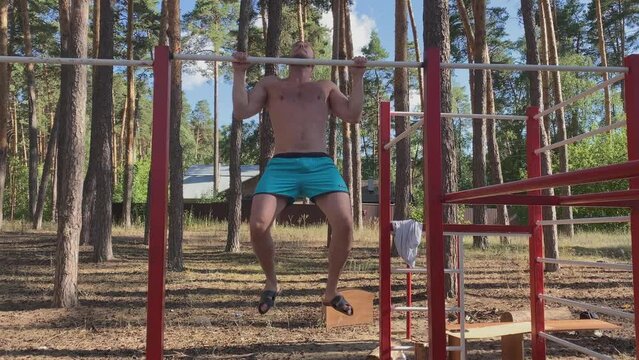 A young athletic man with a naked torso, dressed in blue shorts, does push-ups on the uneven bars at a training ground in the forest. The concept of a zor lifestyle. Summer