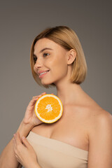 cheerful woman with naked shoulders holding orange and looking away isolated on grey.