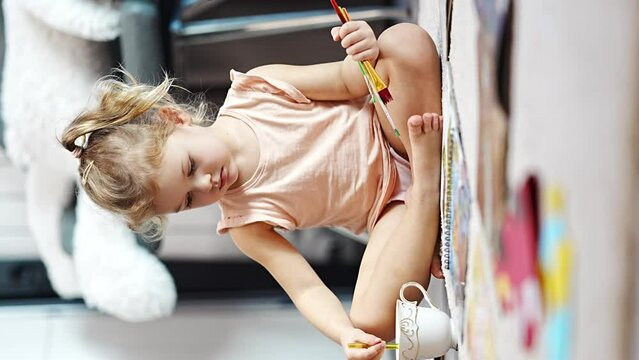 Little girl sits on the floor at home and draws with paints and brushes in a coloring book. Early childhood creativity and education