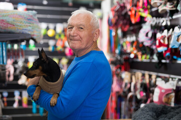 Photo of old man holding miniature pinscher puppy in hands while standing in salesroom of pet shop.