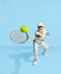 Serving ball. Portrait of handsome senior man in stylish white outfit playing tennis over blue background. Concept of leisure activity, hobby, lifestyle