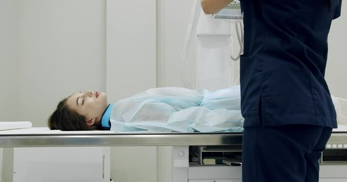 Female patient on X ray machine. Pan left and right view of woman in disposable robe lying on table during belly X ray scanning procedure in modern laboratory.