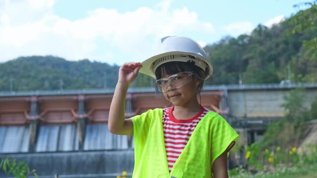 Portrait of a little girl engineer wearing a green vest and white helmet smiling happily on the background of the dam. Concepts of environmental engineering, renewable energy and love of nature and fa