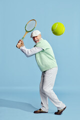 Happy. Portrait of handsome senior man in stylish outfit playing tennis, serving ball over blue background. Concept of leisure activity, hobby, lifestyle