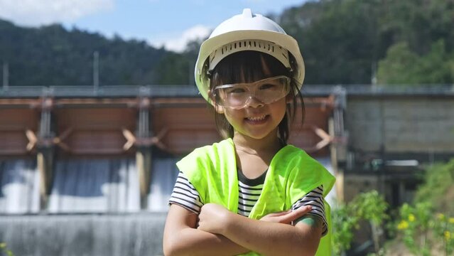 Portrait of a little girl engineer wearing a green vest and white helmet smiling happily on the background of the dam. Concepts of environmental engineering, renewable energy and love of nature and fa