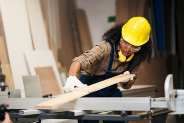 Young attractive black female carpenter wearing safety goggles and hard hat for woodworking in small business workshop. Wood  industry and furniture industry.