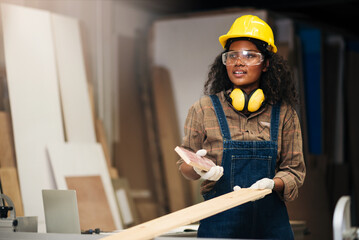 Young attractive black female carpenter wearing safety goggles and hard hat for woodworking in small business workshop. Wood  industry and furniture industry.