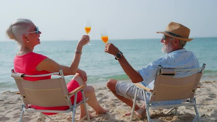 Happy Senior couple drinking orange juice together at tropical beach. Elderly retired husband and wife enjoy summer outdoor lifestyle on beach at holiday. travel and vacation after retirement concept.