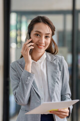 Beautiful Asian businesswoman standing and talking on the phone in office and looking at documents explaining to clients to listen and understand work.