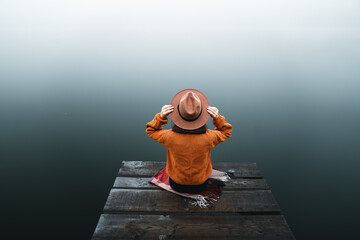 Back view of fashioned young woman standing on wooden dock looking at view on a misty morning. Female hipster with brown hat relaxes on the edge of jetty admiring foggy lake. Wonderful nature getaway