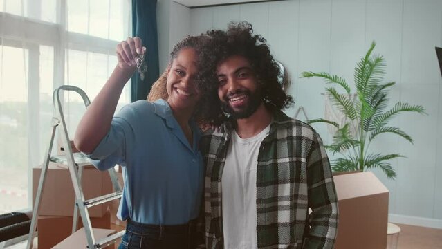 Young happy African American woman and Arabian man with smile show keys to new apartment bought with mortgage and bragging first real estate embracing stand in room near boxes and ladders