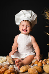  Cheerful little boy sits in cook costume among baking bread rolls on the table on black background
