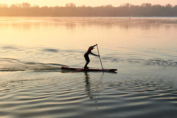 Man rowing on SUP (stand up paddle board) at sunrise in a foggy haze in the Danube river at cold season