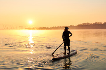 Silhouette of man standing on SUP (stand up paddle board) at sunrise in a foggy haze in the Danube river at cold season