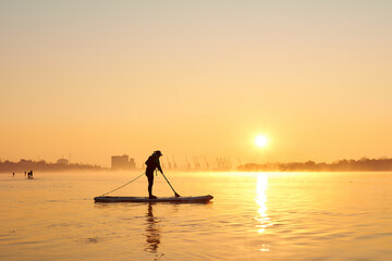 Silhouette of woman standing on SUP (stand up paddle board) at sunrise in a foggy haze in the Danube river at cold season