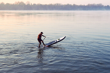 Man rowing on SUP (stand up paddle board) at sunrise in a foggy haze in the Danube river at cold season