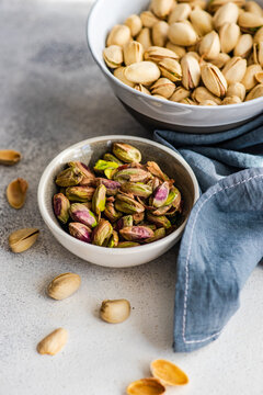 A bowl of pistachio nuts in shells next to a bowl of shelled pistachios