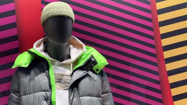Mannequin in a down jacket, hoodie and a knitted hat stands in the window of a clothing store. View through the glass