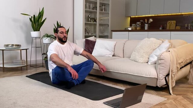 Mid adult bearded man using laptop meditate at home. Tranquil male meditating by following online yoga classes via computer indoors. Calm guy practicing contemplation by video tutorial domestic room