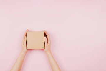 Top view to female hands holding small rectangle brown cardboard box on pastel pink background....