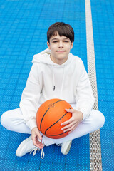 Close up. Teen athlete with basketball on background of blue sports court. Sport and hobby concept. White tracksuit.